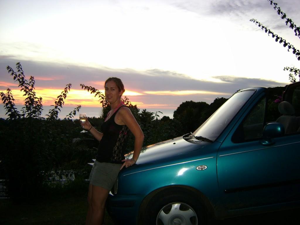 young woman leaning against bonnet of green car at sunset with glass of white wine in her right hand