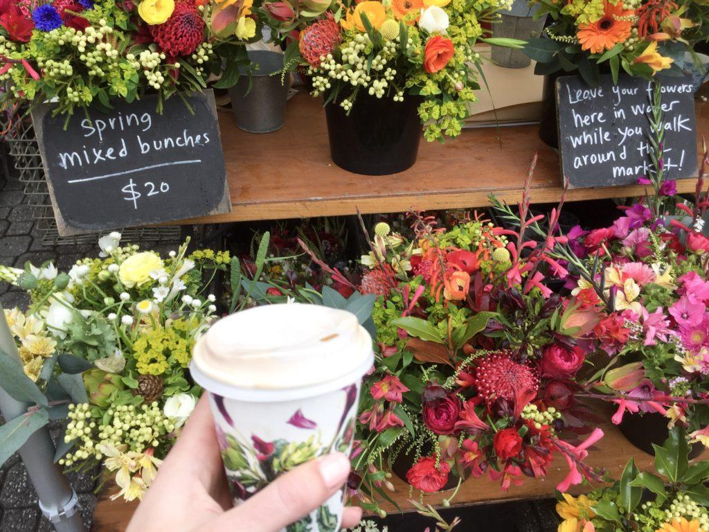 hand holding takeaway coffee cup in front of flowers sold at market
