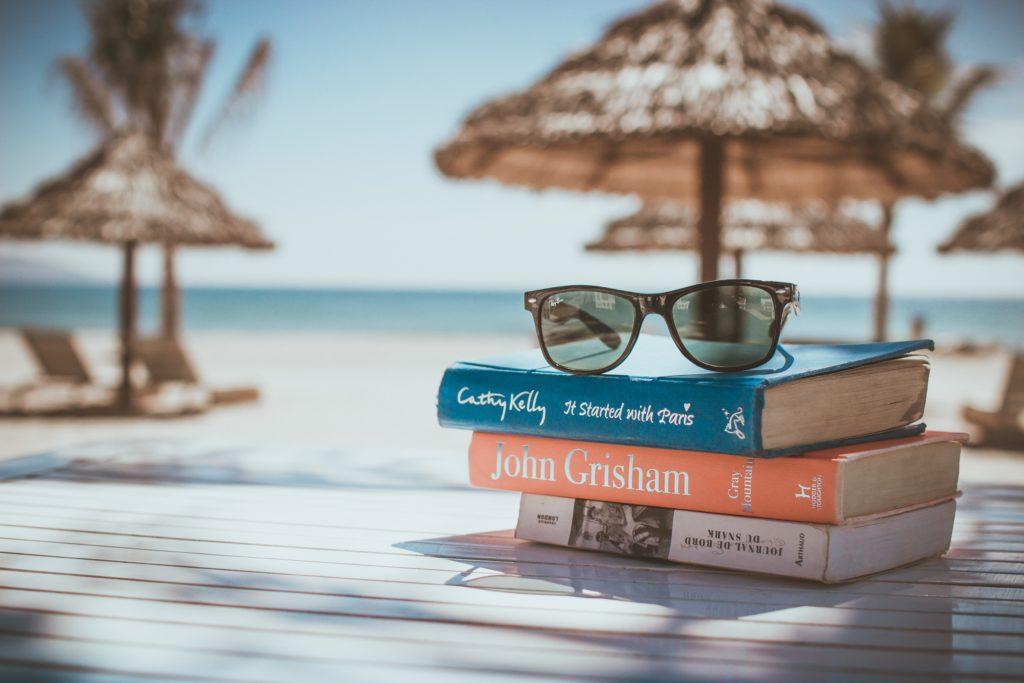 books to read on holiday stacked under sunglasses on beach