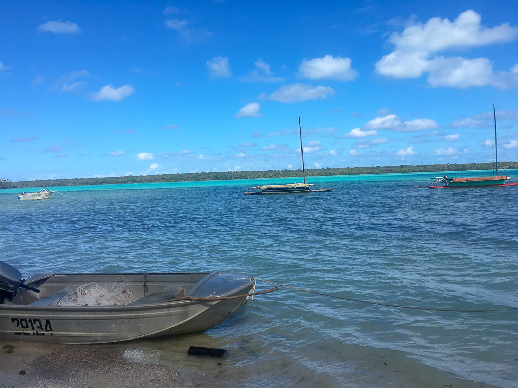 three handmade boats on blue water with a speed boat grounded on sand in foreground