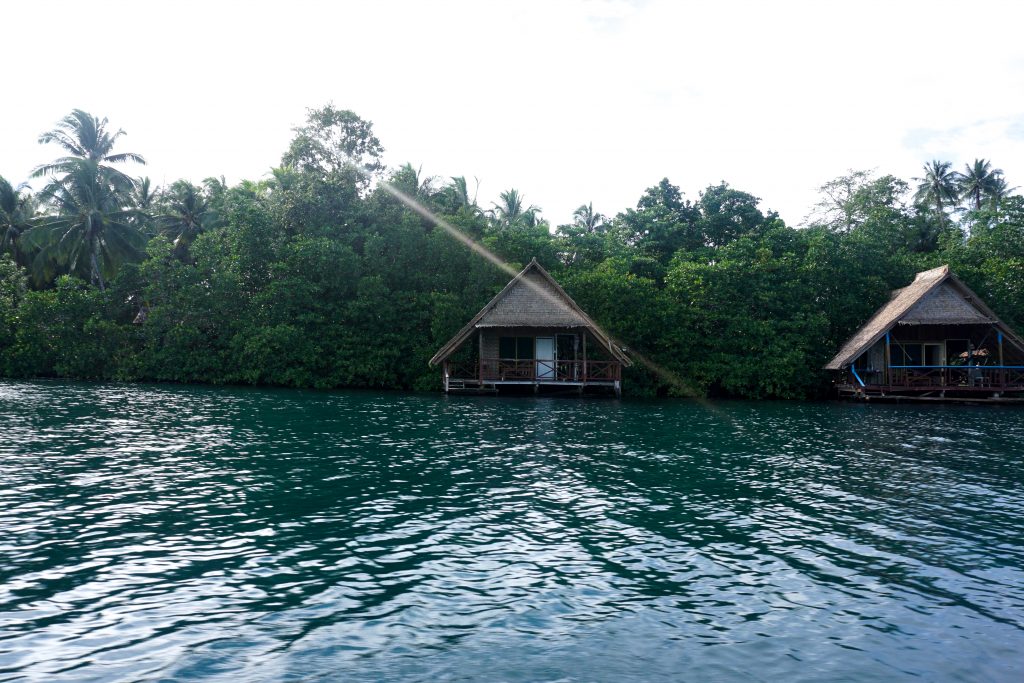 two simple bungalows at the edge of an island viewed over water from a boat