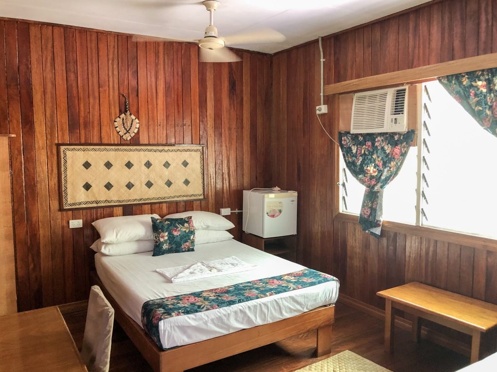 double bed with white linen in wooden paneled motel room with ceiling fan and floral accents