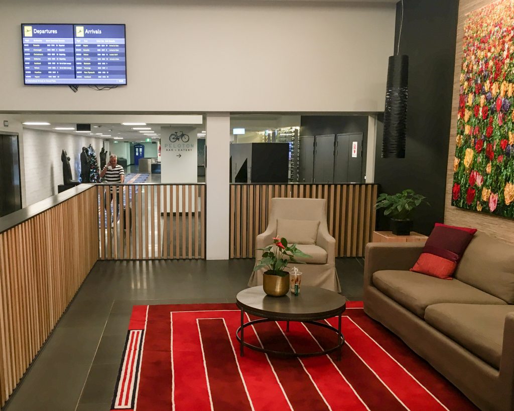 Rydges Wellington Airport lounge area with tan couch, table, red carpet and departures board