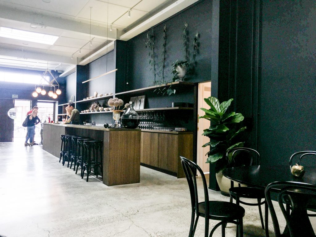 long bar in gin distillery with concrete floor, two greet plants, a table with four chairs, barstools and glasses