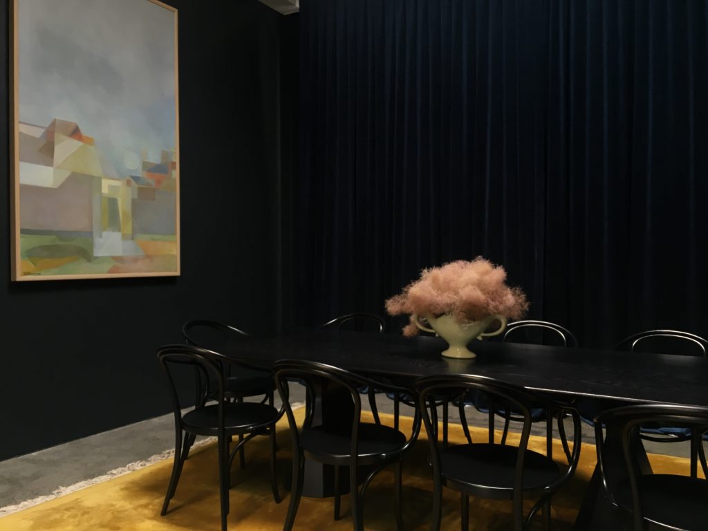 Dark room with long black table, 9 chairs, pink floral centerpiece, large artwork on wall and yellow carpet