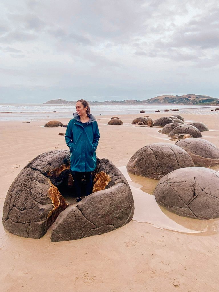young woman in green raincoat standing in boulder on Moeraki Beach New Zealand