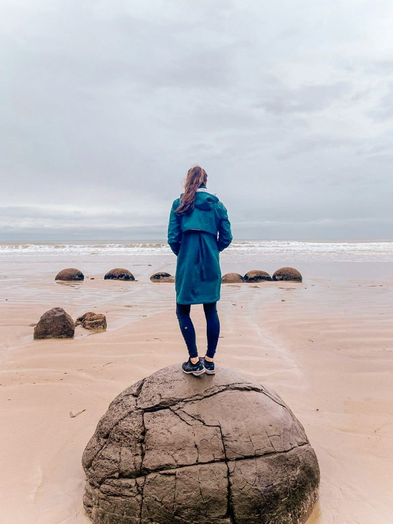 young woman in green raincoat standing on boulder on Koekoehe Beach New Zealand