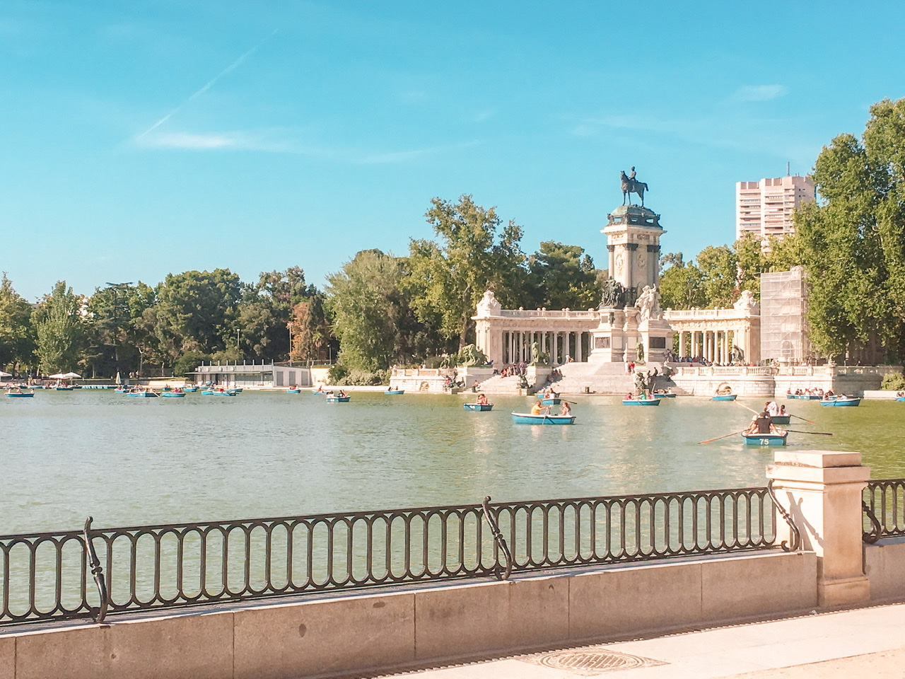 numberous blue boats with people rowing on blue lake in front of statue and monument on a Sunny day at Retiro Park in Madrid