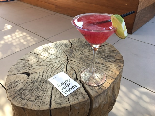 red cocktail with lime wedge and short black straw on tree stump table next to room card which says Enjoy your room