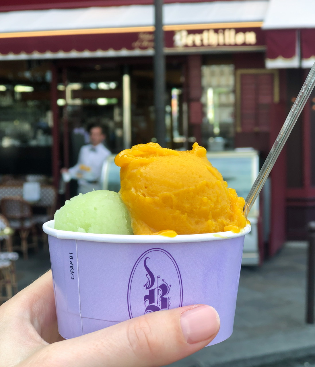 Scoops of mango and green apple sorbet in a purple cup held in hand in front of Berthillon store front