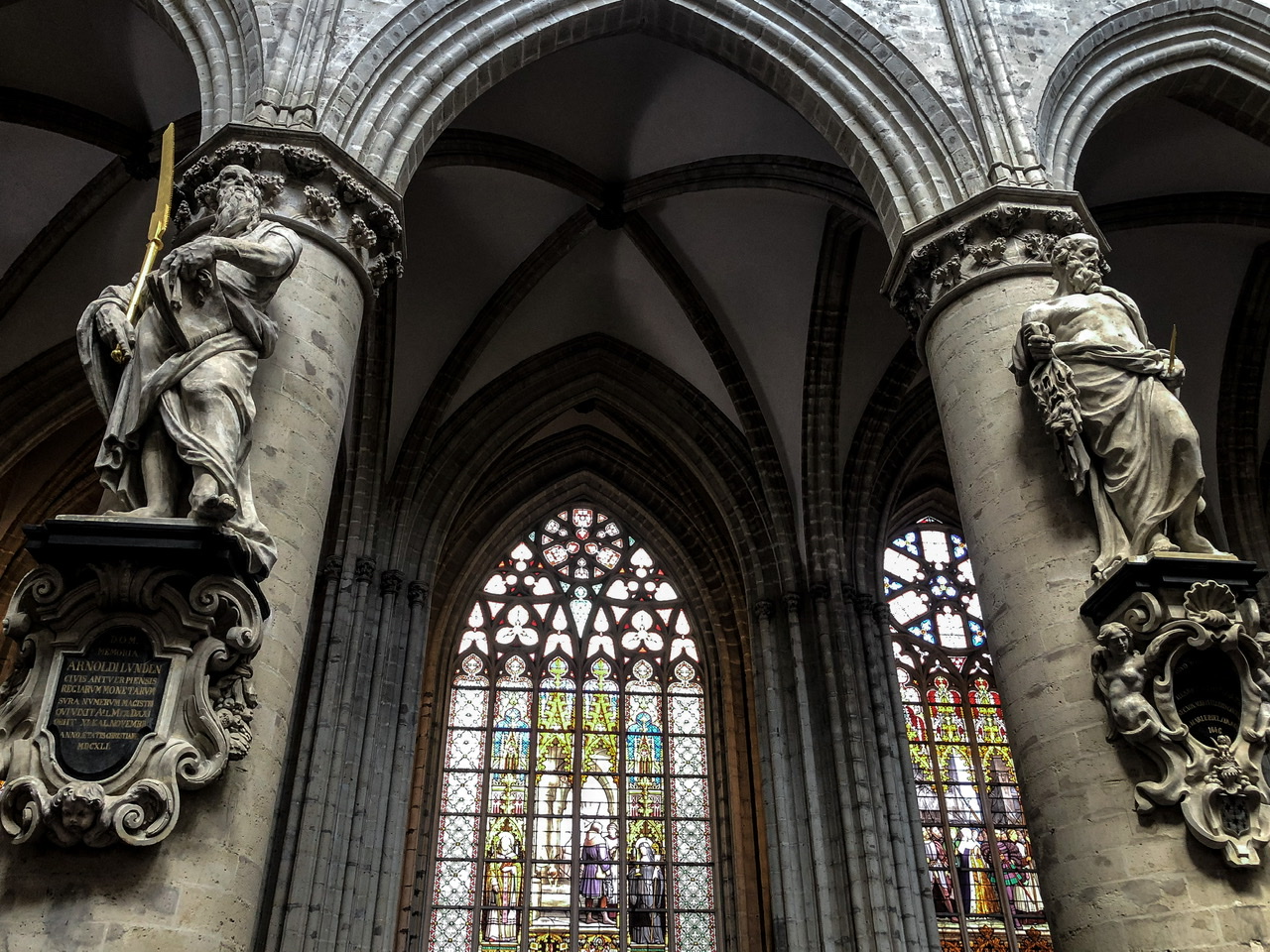 Two statues of apostles on pillars with a stained glass window between inside St Michael and St Gudula Cathedral in Brussels