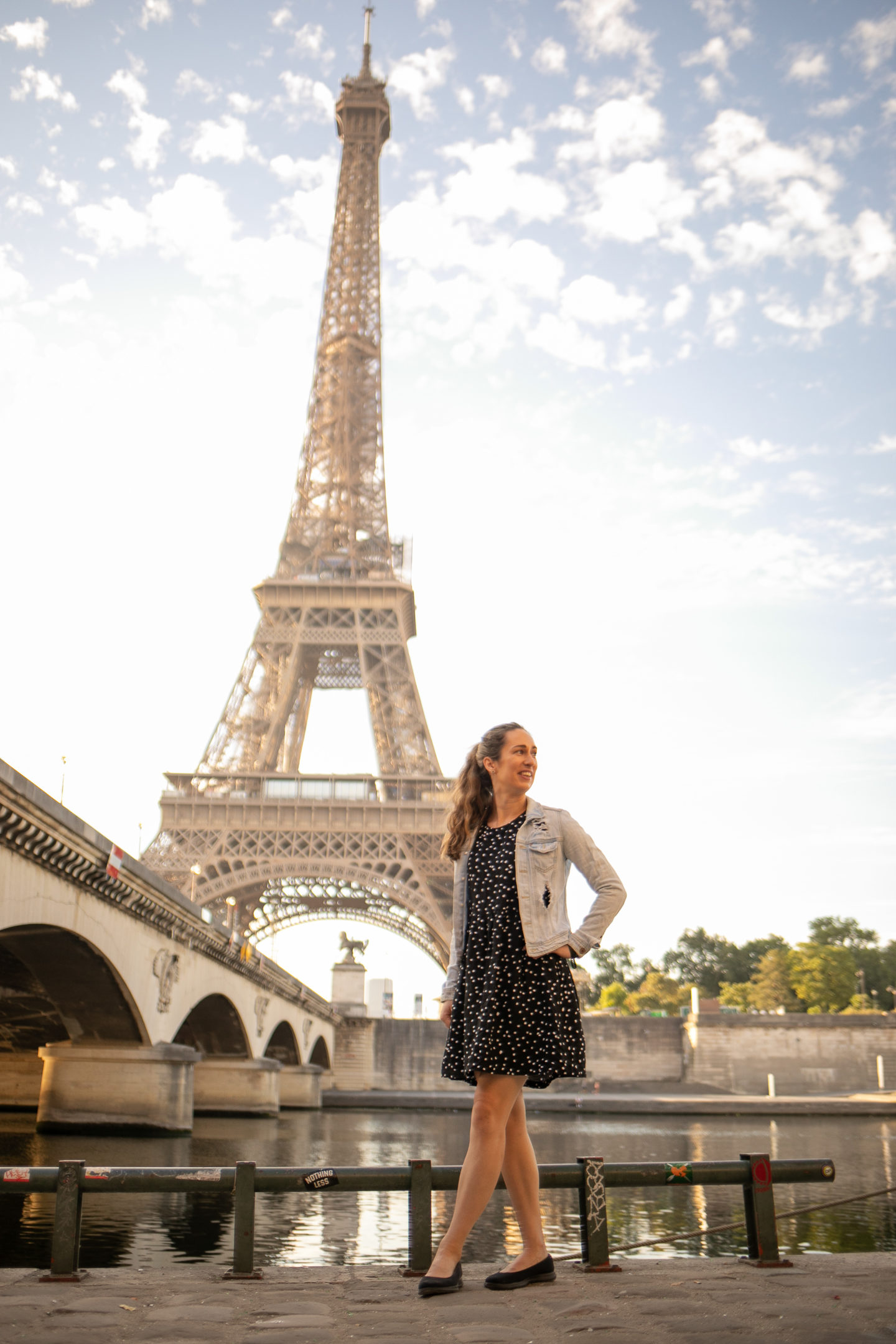 Young woman in blue dress and denim jacket standing in front of River Siene with Eiffel Tower in background