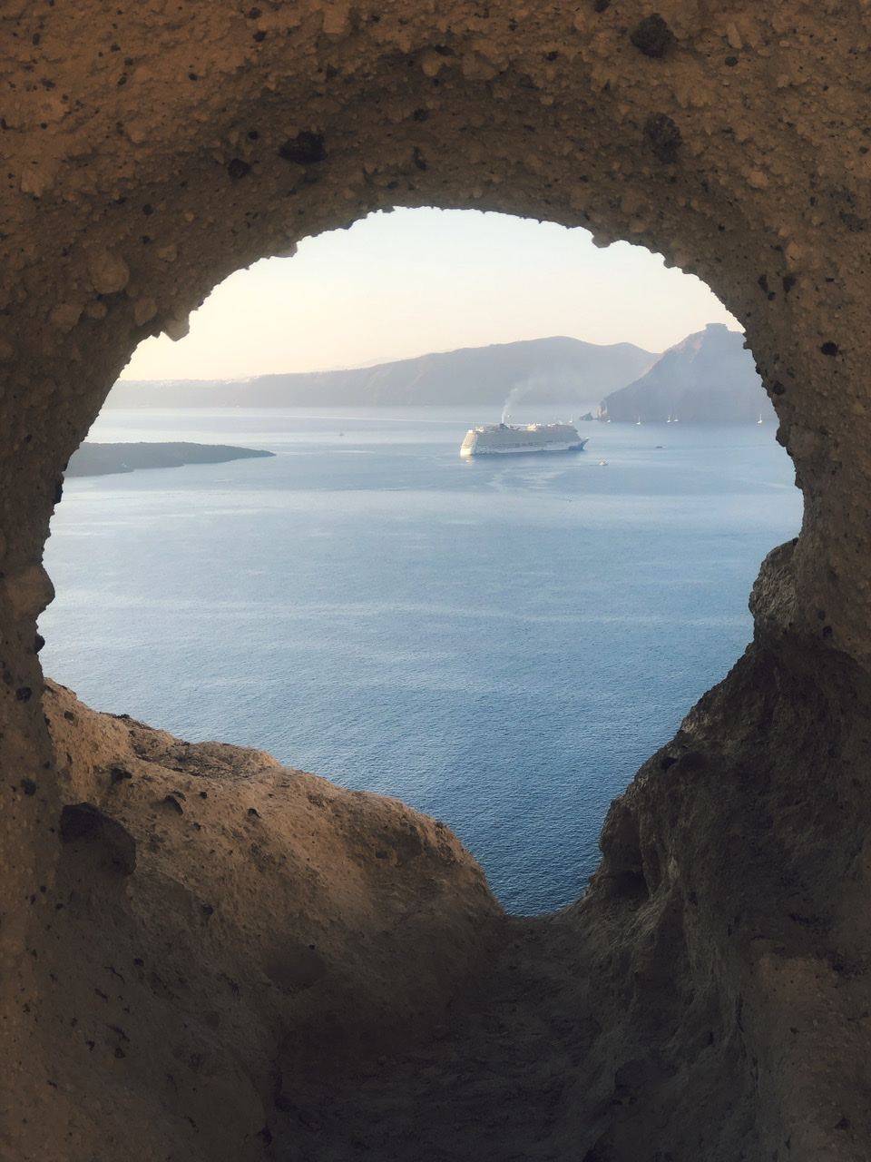 cruise ship at sea with volcanic greek islands in background pictured through heart shaped hole in rock in santorini