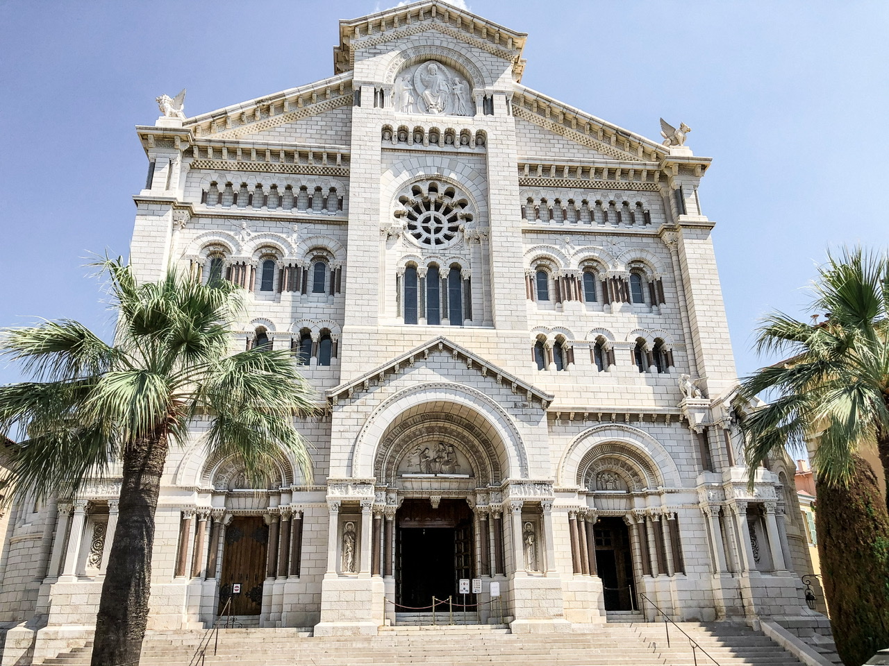 Exterior of Cathédrale de Monaco, a white gothic era cathedral with steps leading up to it and palm trees either side