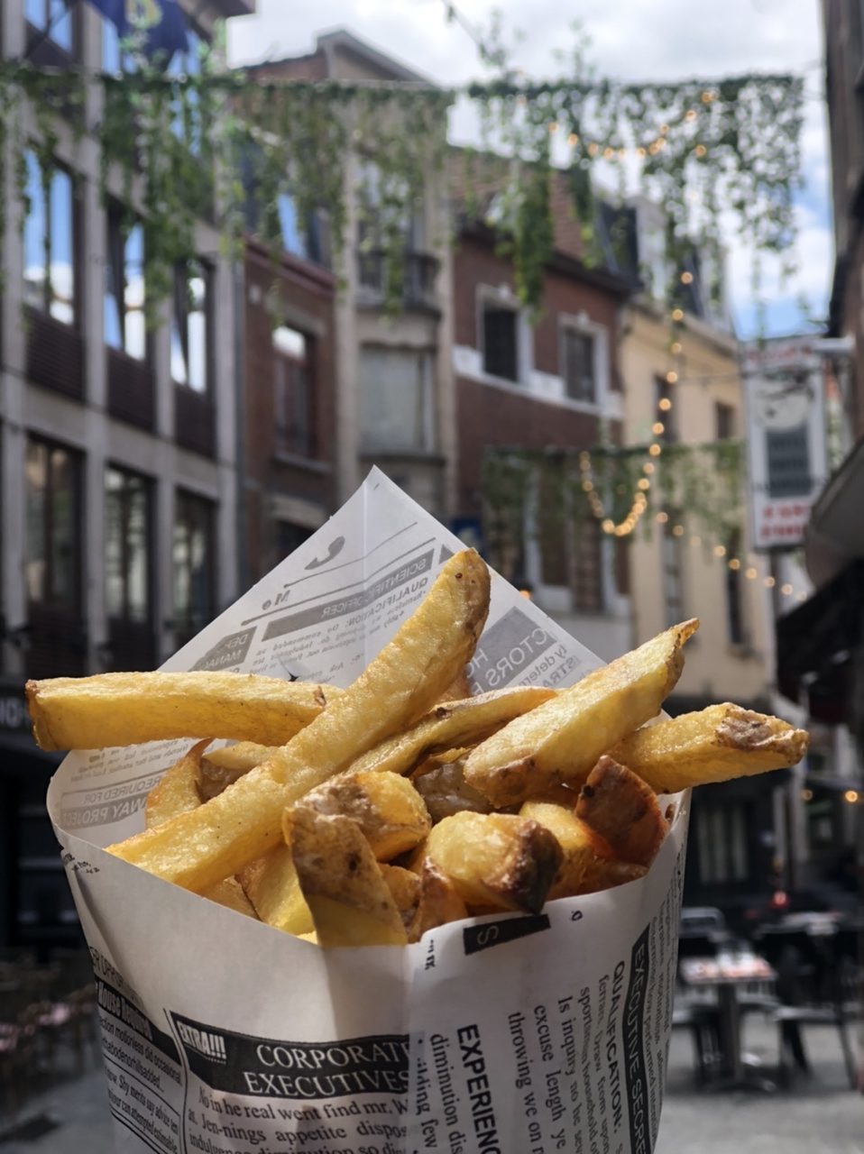 Fries in funnel formed out of printed paper designed to look like newspaper with held up with four tall Brussels buildings in background