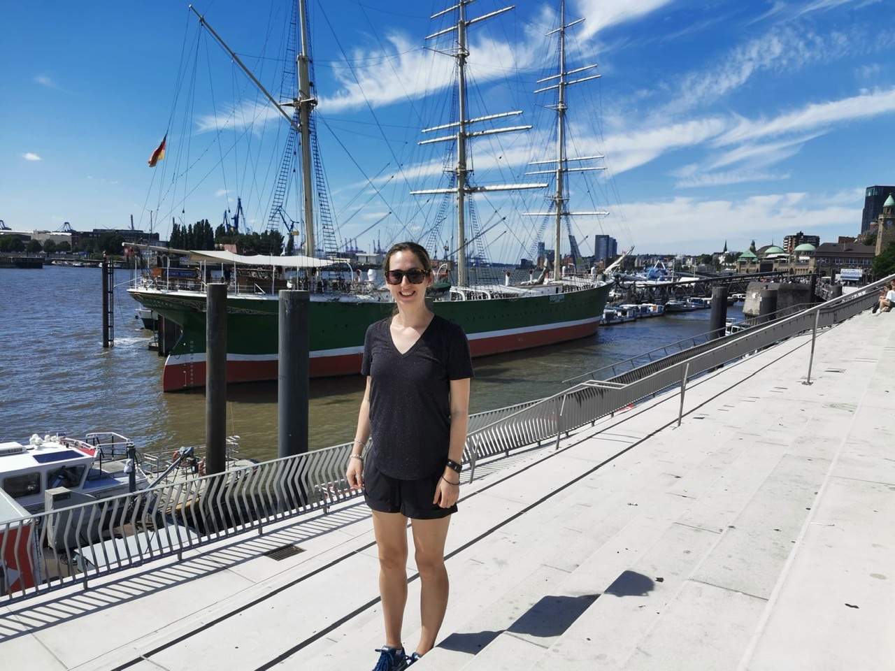 Young woman dressed in black shorts, t-shirt and sunglasses in front of tall ship at Hamburg Waterfront