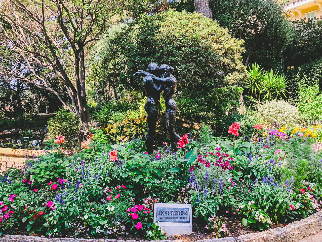 Statue of couple embracing surrounded by green fauna with pink and purple flowers in Les Jardins Saint-Martin