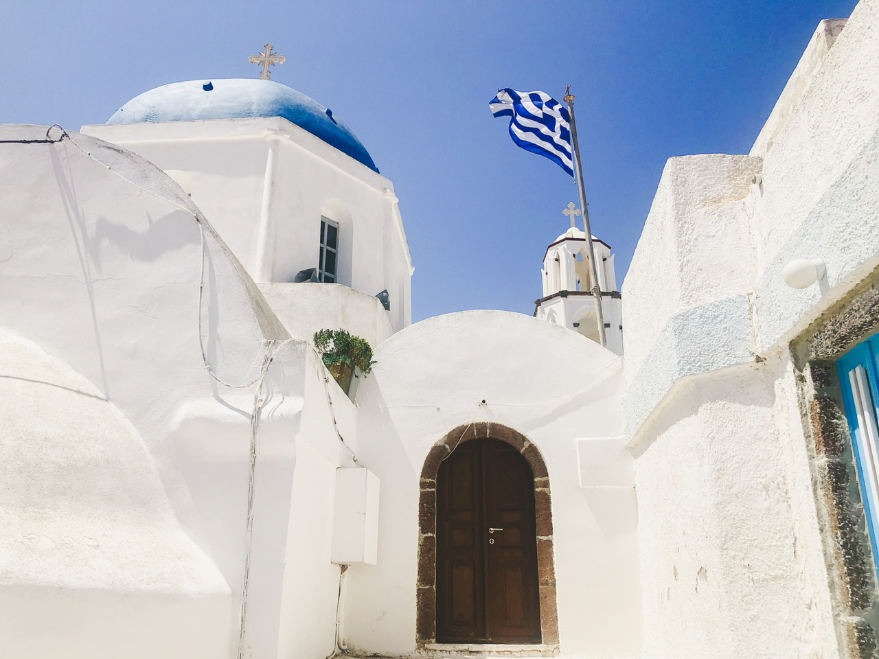 White church with blue dome and brown wooden door with greek flag flying in Santorini