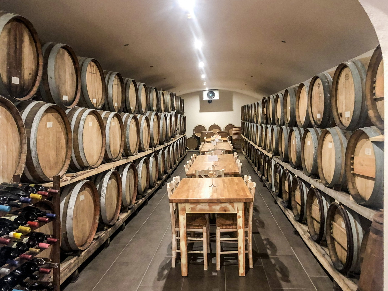 rows of 10+ wine barrels stacked three high on either side of a cellar with three tables each with four chairs running length of the centre of the room. Wine rack holding many bottles of wine in left-hand front corner