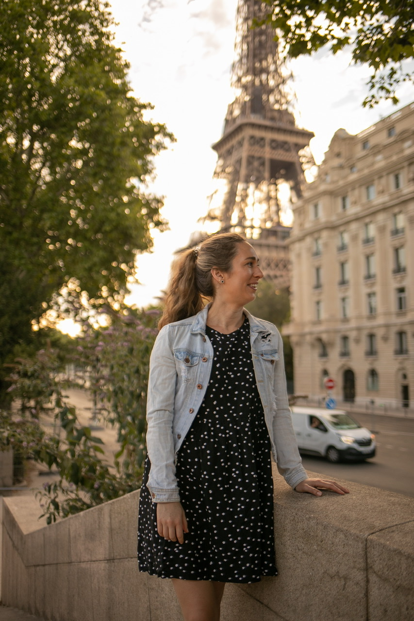 Young woman in blue dress and denim jacket leaning on concrete edge with a street, white van, Parisian building and Eiffel Tower in background