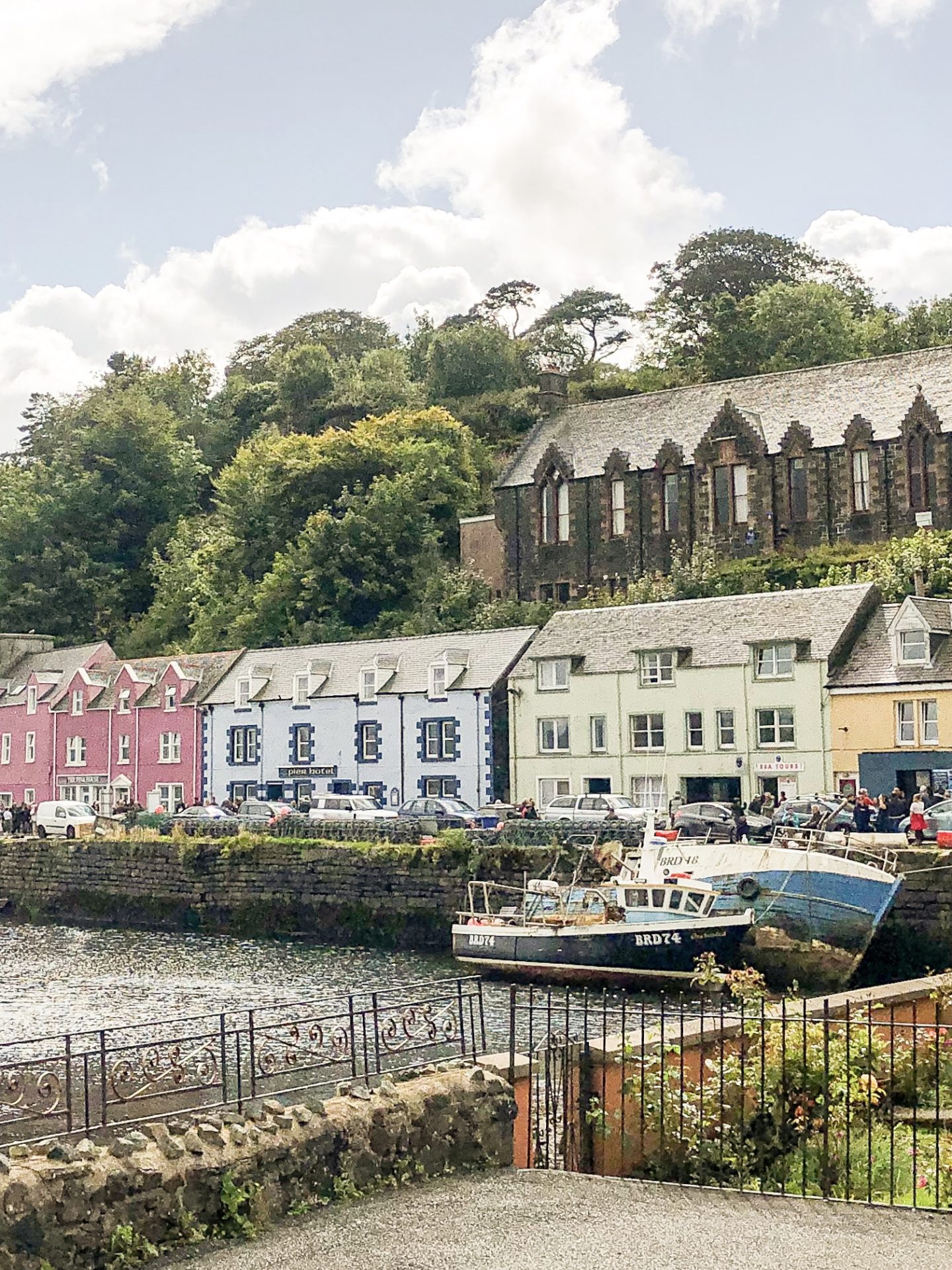 Wharf with two old ships moored in right-hand corner. Multi-story port buildings in pink, blue, green and orange with an older brown church building behind them on green hill.