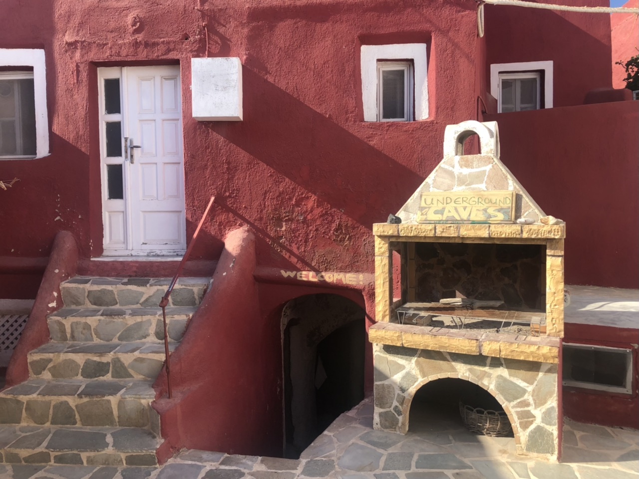 Red concrete building with three windows and stone steps leading up to white door. Outside there is an outdoor oven and signs saying "underground caves" and "welcome".