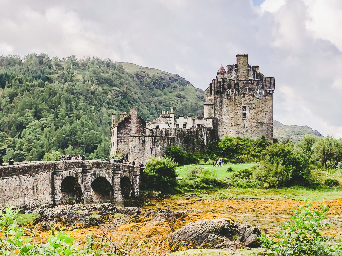 Exterior of Eileen Donan Castle with stone footbridge to left, green covered hills in background to left and mossy green terrain to right in front of the castle