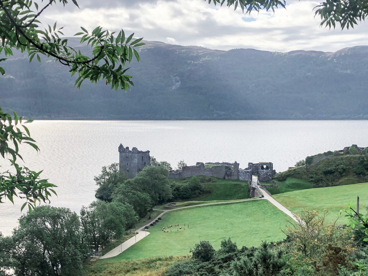 Exterior of Urquhart Castle in distance behind green fiends on edge of Loch Ness in Scotland. Green hills seen on far side of the lake.