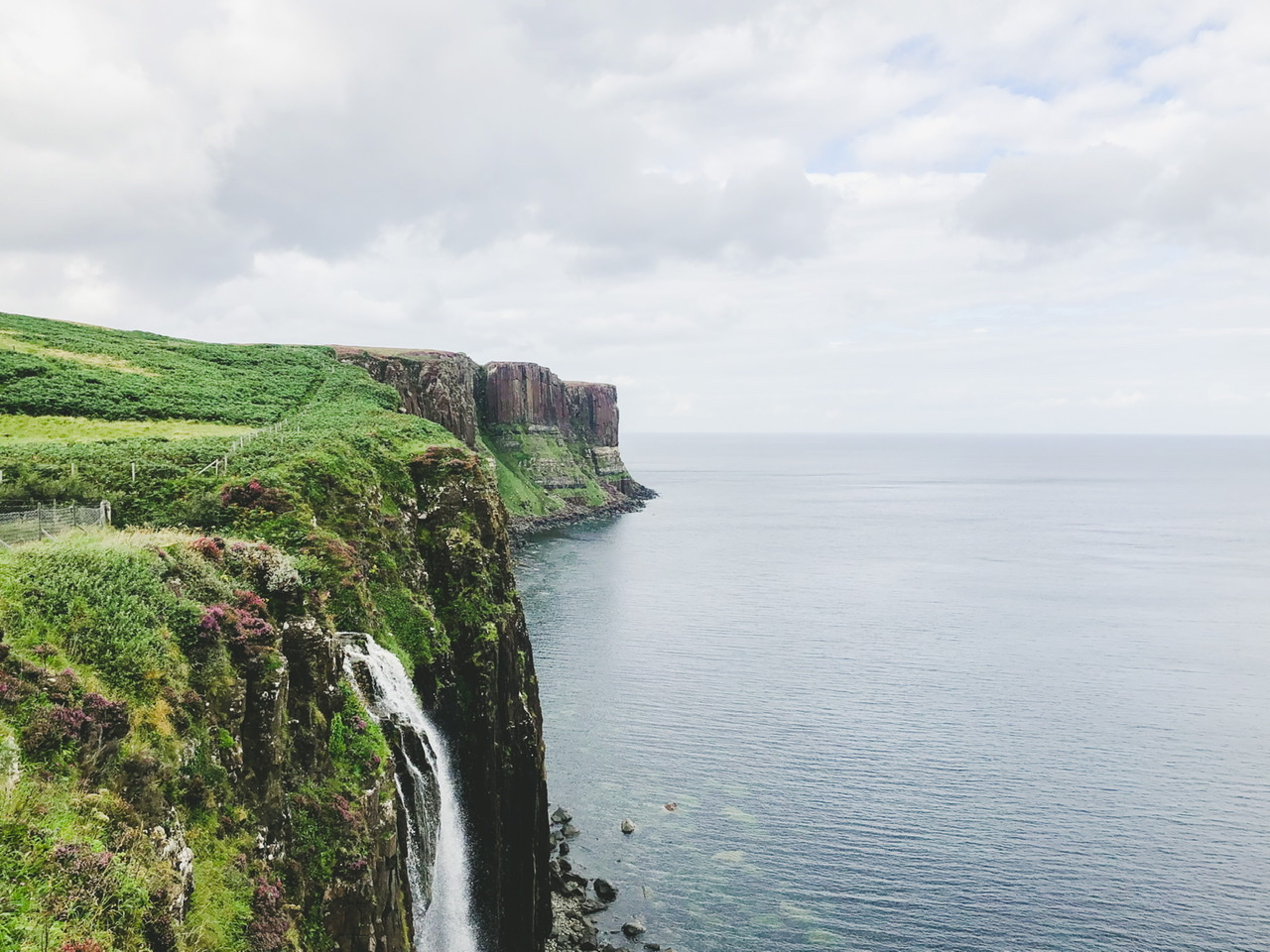 Green covered cliffs and Mealt Falls on the right fall away into rocky ocean on a cloudy day in Scotland