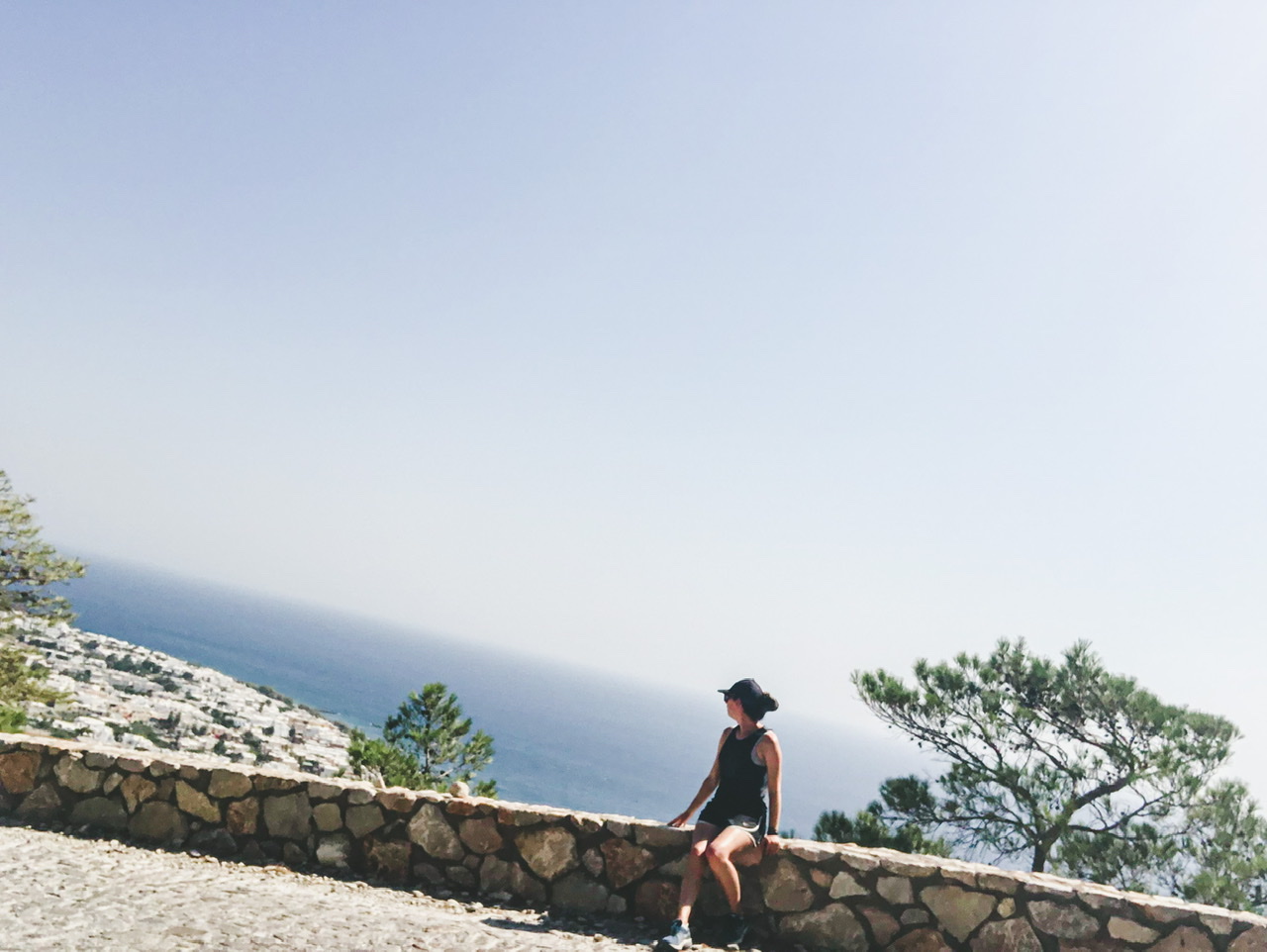 Young woman sitting on wall on side of road up to Ancient Thera with blue sea and buildings of Kamari below