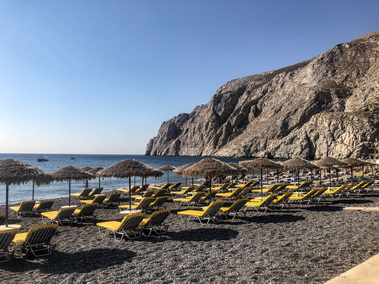 Volcanic black stoney beach covered in yellow sunloungers with woven sun umbrellas, two small boats in ocean and rocky cliffs to right
