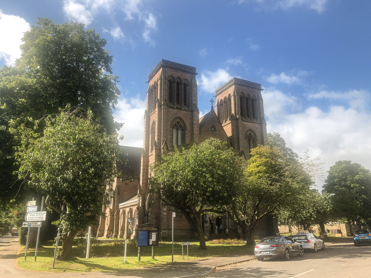Exterior shot of brown sandstone cathedral with no spires on a grassy block with leafy green trees to the right and front