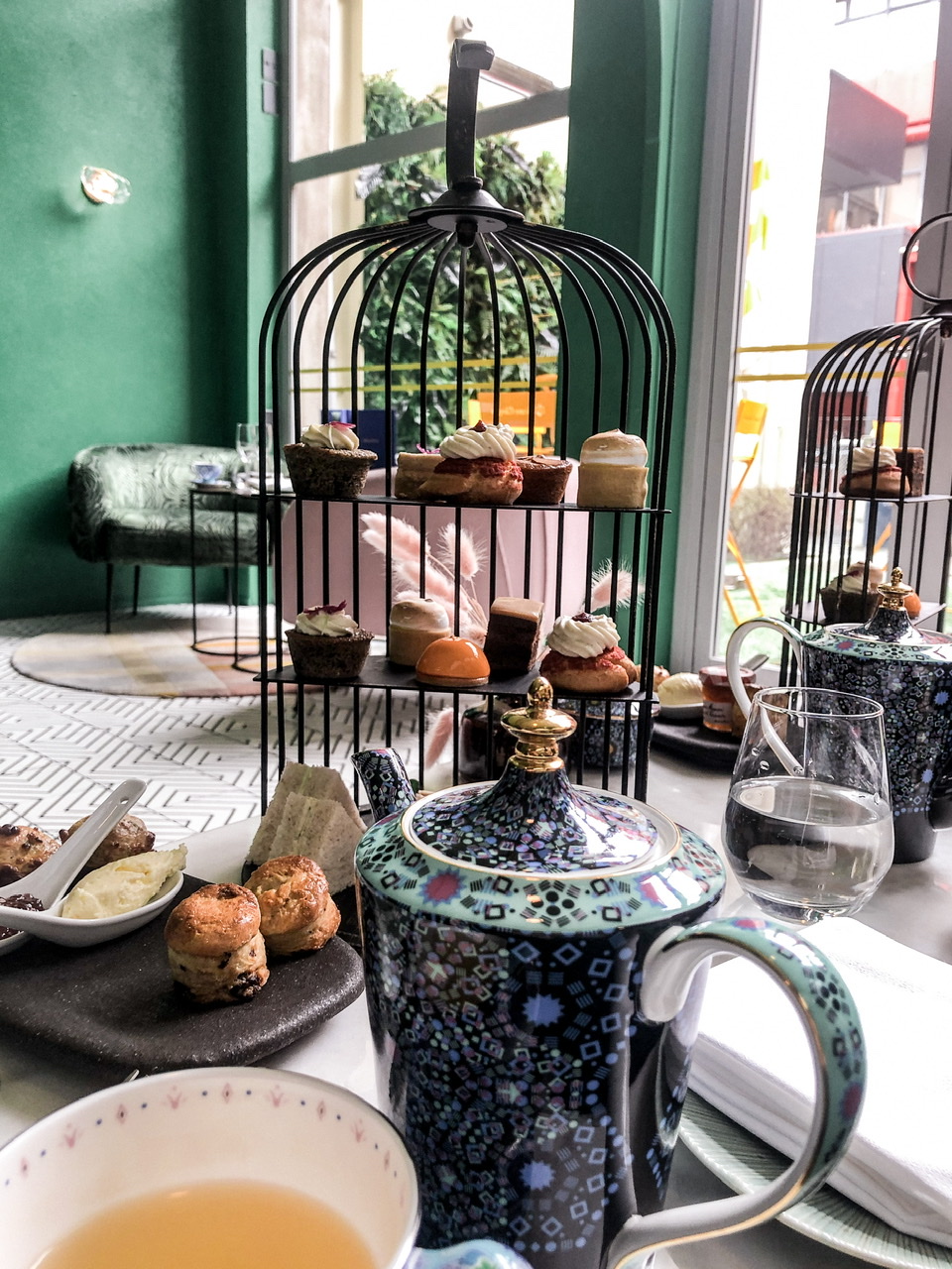 Three tiered high tea stand with plate of four small scones with cream, talk blue flowery tea pot and poured cup of herbal tea in foreground, green painted wall, window and couch in background
