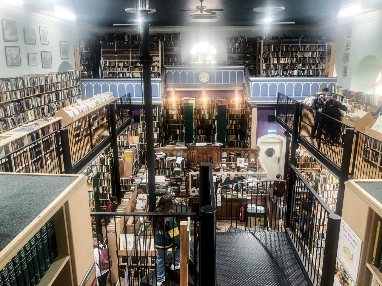 Interior of old bookshop inside a repurposed church. The second floor is formed of balconies leaving the ground floor and log fire chimney visible in the centre. Two people browsing upstairs, two down, and a young woman is climbing up circular metal staircase in front centre.