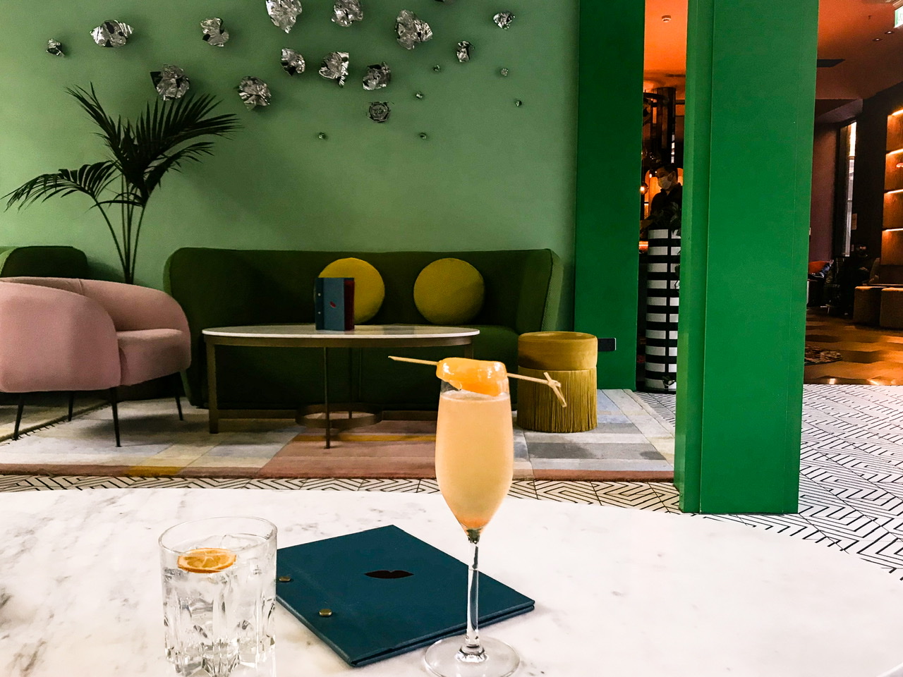 Room with bright green walls and art deco couch with champagne cocktail decorated with a lychee and glass of water containing a lemon slice placed next to closed bar menu on table