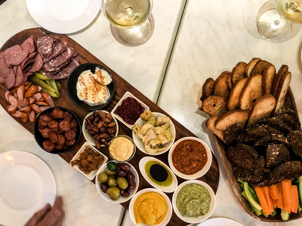 Platter of cured meats, dips and olives alongside a basket of breads and crudités with two glasses of chardonnay and three side plates on white table