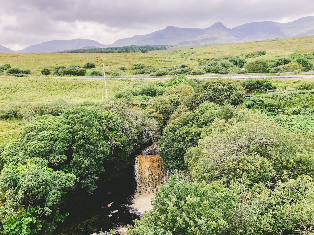 Muddy waterfall in centre with greenery either side, rolling mountains in background and main road passing through centre of background