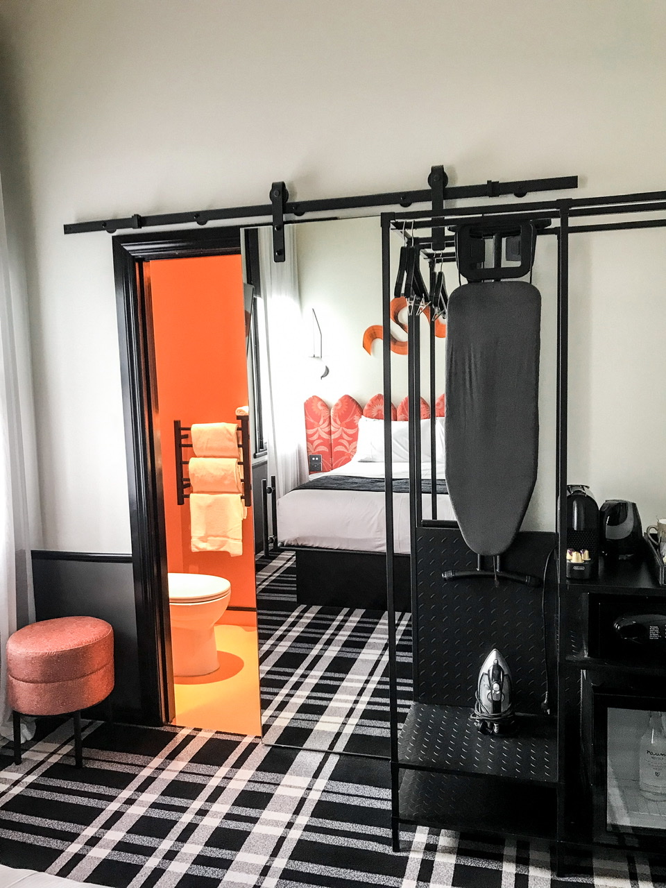 Toilet and three white towels visible in orange painted bathroom behind ajar sliding door with hotel bed visible in mirror on sliding door. Black and white tartan carpet.