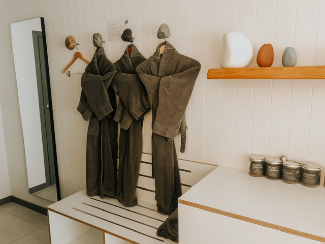 Three grey Bathrobes hanging on a white wall between a full length mirror and a floating wooden shelf with three coloured vases above a bench with four jars of tea on it.