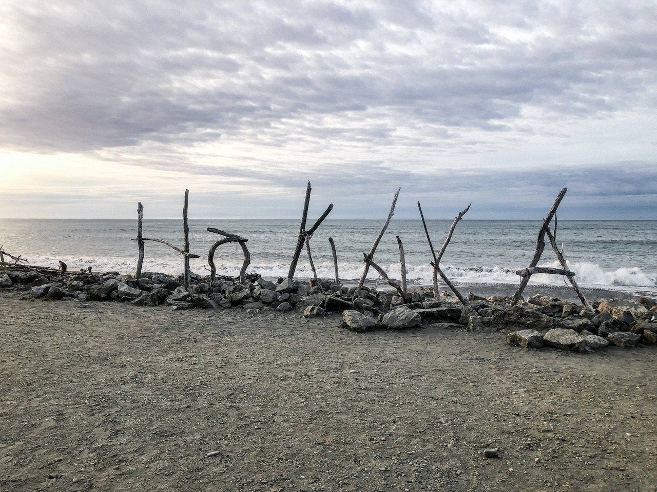 “Hokitika” spelt out in driftwood standing vertically on a beach with ocean behind it