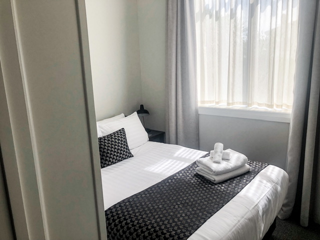 Double bed in apartment bedroom with white linen and black blanket across it and matching black cushion. Window with lightly coloured curtains and lace curtain. Black bedside table and lamp in top right corner of bedroom.