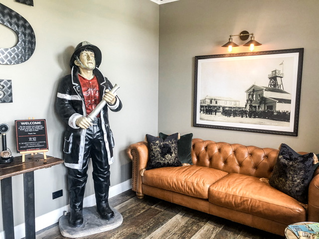 foyer at firestation boutique accommodation hokitika with a mannequin fireman, large brown leacher couch under a historic photo of the firestation.