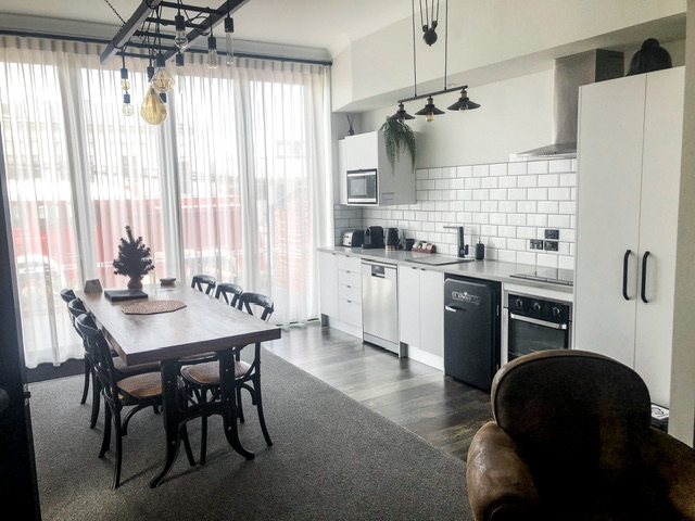 Open plan kitchen and dining area in Hokitika Firestation apartment. On the right is bench with oven, dishwasher and fridge underneath. A wooden take in centre with thee chairs each side and a single leather armchare in foreground. Back wall is full length glass covered by lace curtains. An old fire truck is visible outside.