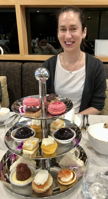 Siver three-tier high tea stand with eight savory and 12 sweet items. A young woman in a white tshirt and grey cardigan sits smiling behind it.