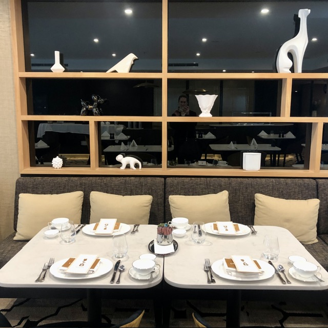 Table set for four people with white plates with menus on top of them, each has a bone china cup and saucer and sugar bowl beside them. Behind the table is soft grey seating with 4 creme cushions and white china ornaments sit on three shelves above.