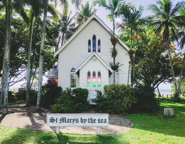 White church with green and pink stained glass windows and sign saying “St Marys by the sea” the church is surrounded by green grass and tall palm trees, the ocean is visible behind the church.