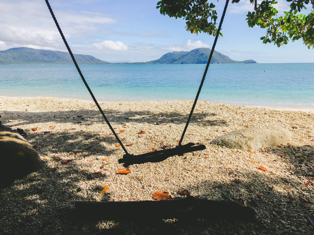 Beach swing made out of driftwood and rope on an island with white sandy beach, sea and far green land visible