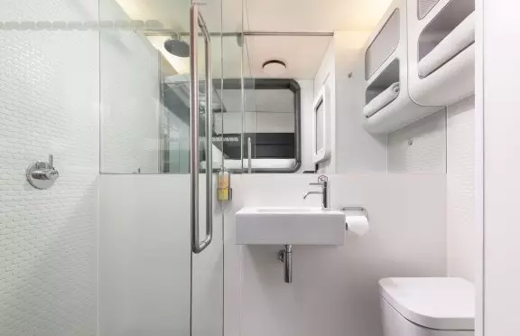 Compact white YotelAir hotel bathroom with toilet on right with shelf with towel above it, mirror and basin straight ahead and shower control on left. There is a glass sliding door in the foreground.