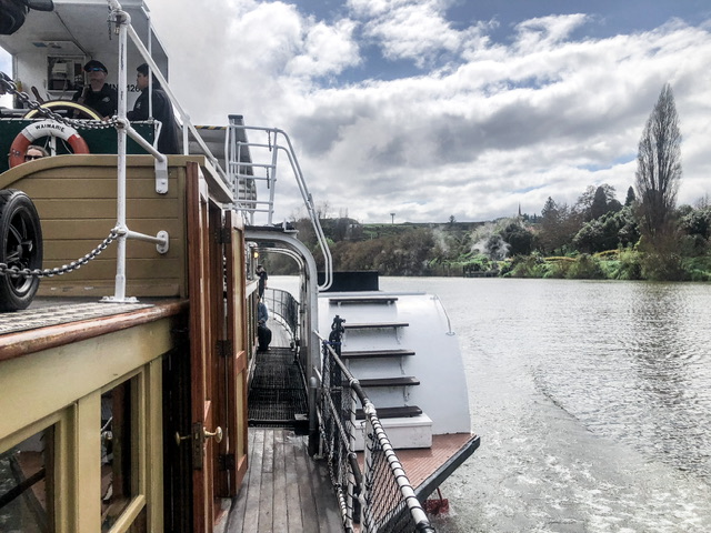 Paddle Steamer Waimarie on the Whanganui River on a sunny day, A foliage covered riverbank is visible on the right. You can see the ship's wheel with two staff operating on the left.