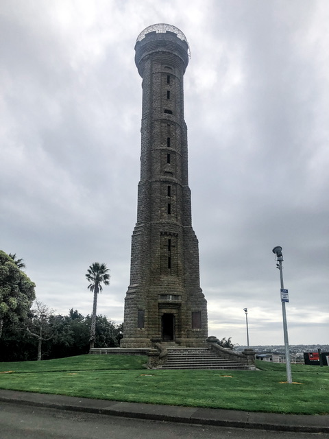 A 33 metre tower of remembrance built out of fossilised shell rock pictured from a distance. It is surrounded by lush green lawn, two tall spotlights on the right and trees on the left.